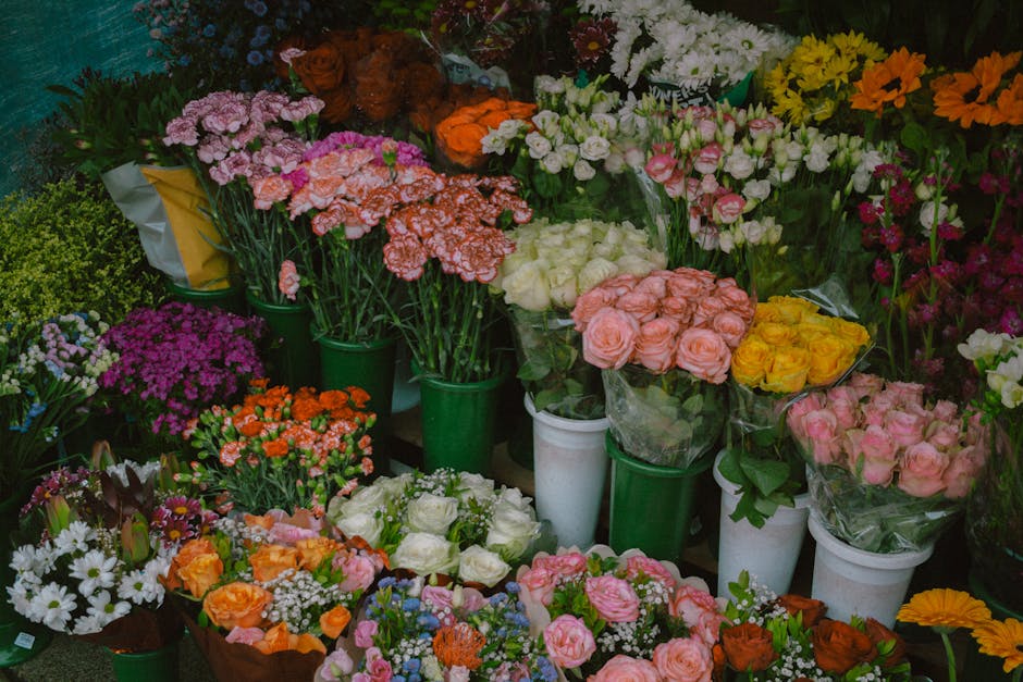 A colorful display of fresh flower bouquets and potted arrangements arranged in green and white containers, featuring a variety of flowers including roses in soft pink, yellow, and white, as well as c