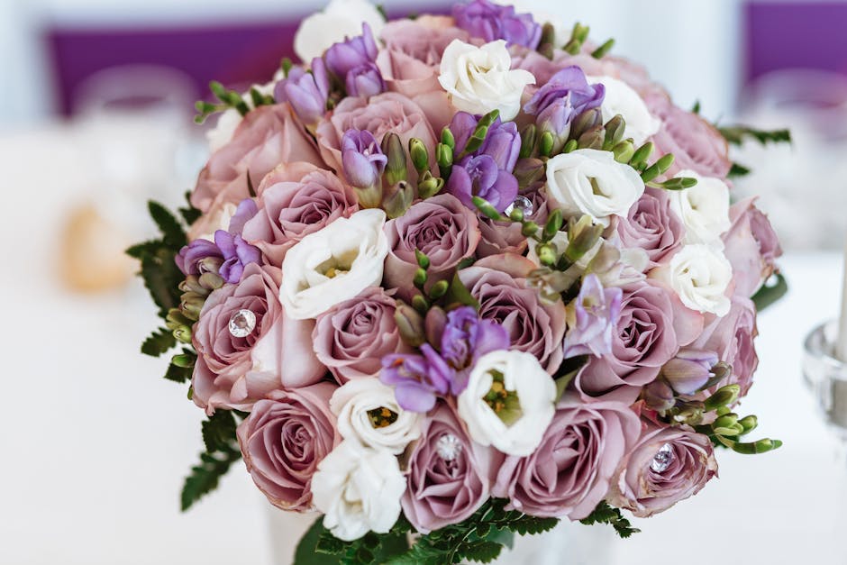 A close-up of a floral arrangement featuring a round bouquet of fresh pink roses, white lisianthus, and purple freesias, accented with small green foliage and a few decorative clear gemstones. The flo