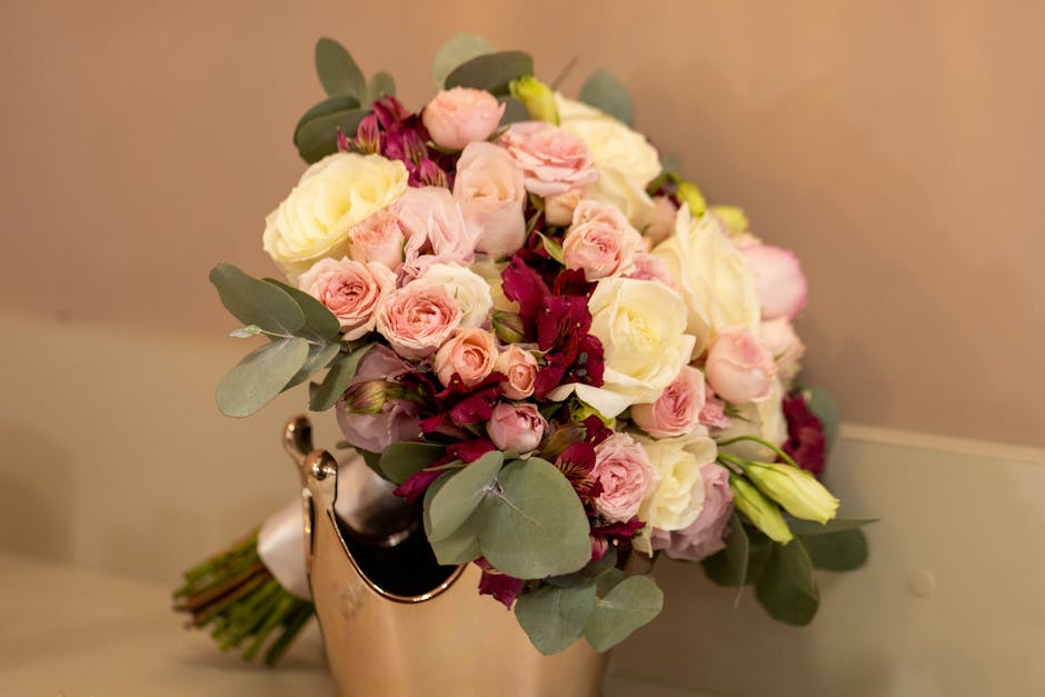 A fresh bouquet arrangement featuring predominantly white and soft pink roses, complemented by deep burgundy alstroemeria and sprigs of eucalyptus with round, silvery-green leaves. The bouquet is arti